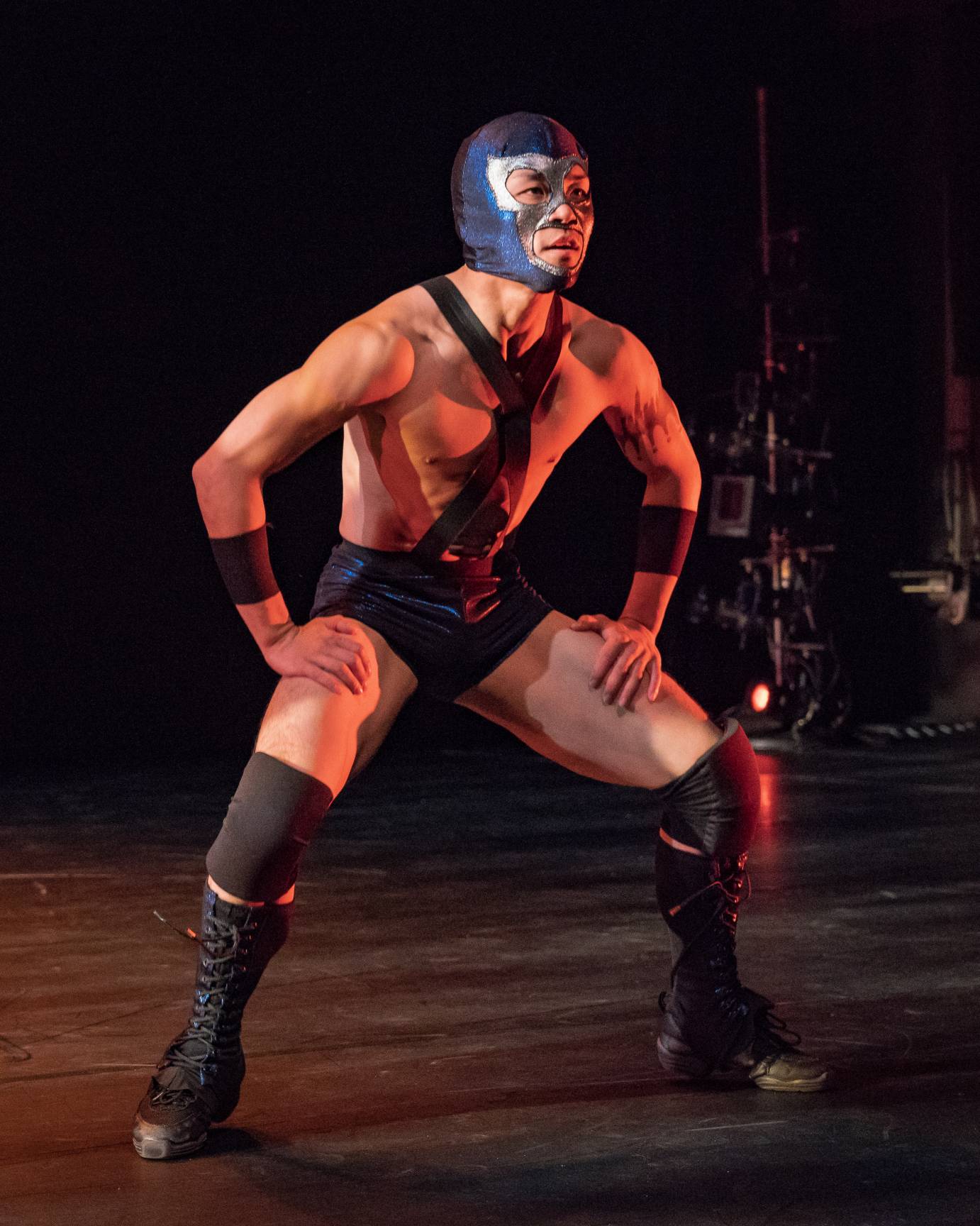 A man in a wrestling mask assumes a squat position looking out to the audience
